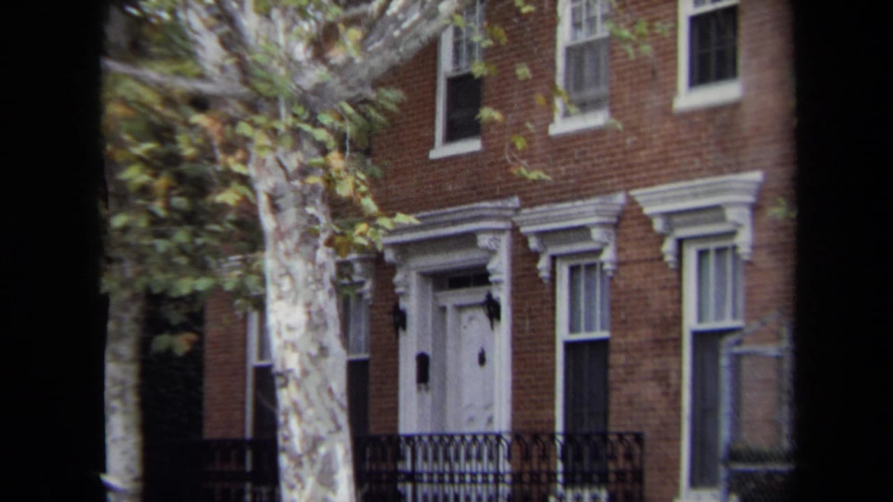 1970 Annapolis home-movie frame showing a brick townhouse facade, white door, black iron fence, and autumn tree beside the street.