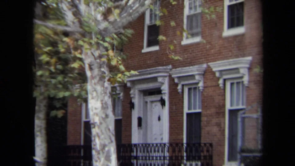 1970 Annapolis home-movie frame showing a brick townhouse facade, white door, black iron fence, and autumn tree beside the street.
