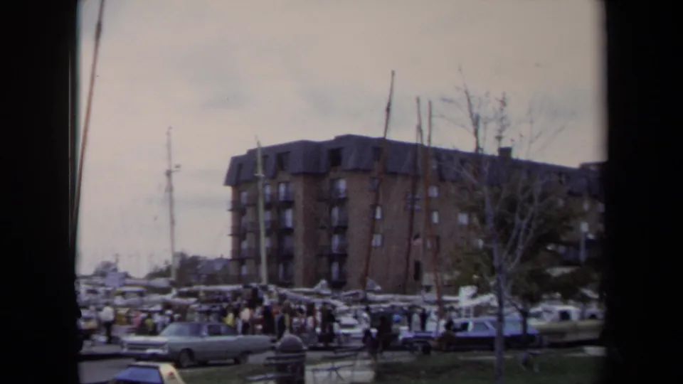 1970 Annapolis marina view with sailboat masts, parked cars, distant visitors, bare trees, and a large brick apartment building.