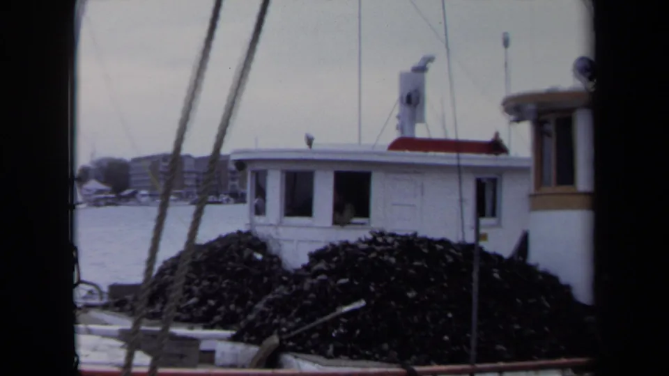 A 1970 Annapolis dockside frame shows a white cabin and large dark shellfish piles, with harbor buildings in the distance.