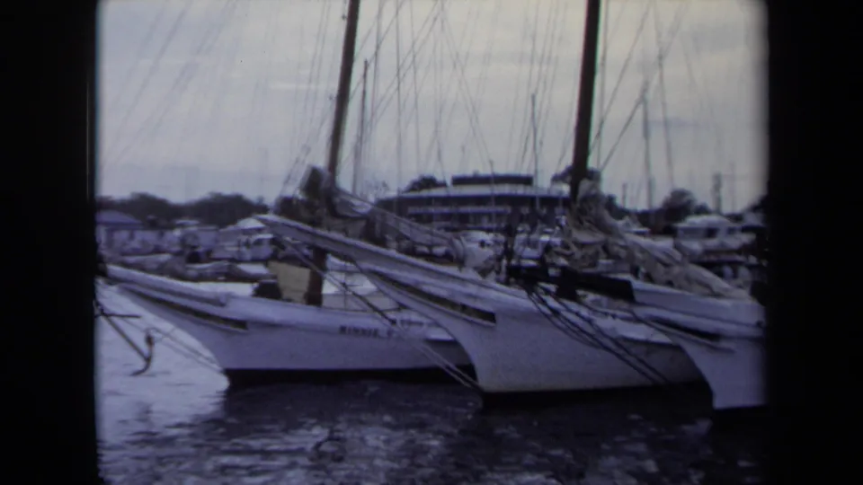 1970 Annapolis marina shot with neighboring white sailboats at their slips, folded sails, mast rigging, rippled water, and cars along the shore.