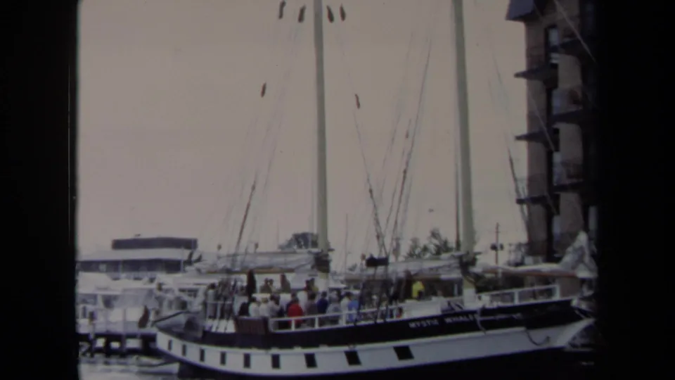 The Mystic Whaler sits at an Annapolis marina with people on deck, tall masts above, and a waterfront building at the right edge.