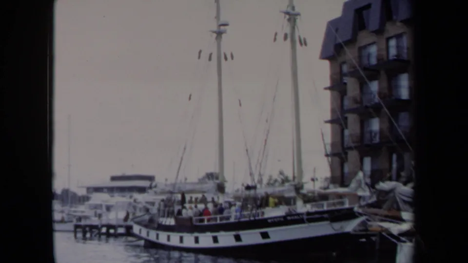 1970 Annapolis harbor view with docked sailboats, tall masts, and a black-and-white boat near waterfront buildings.
