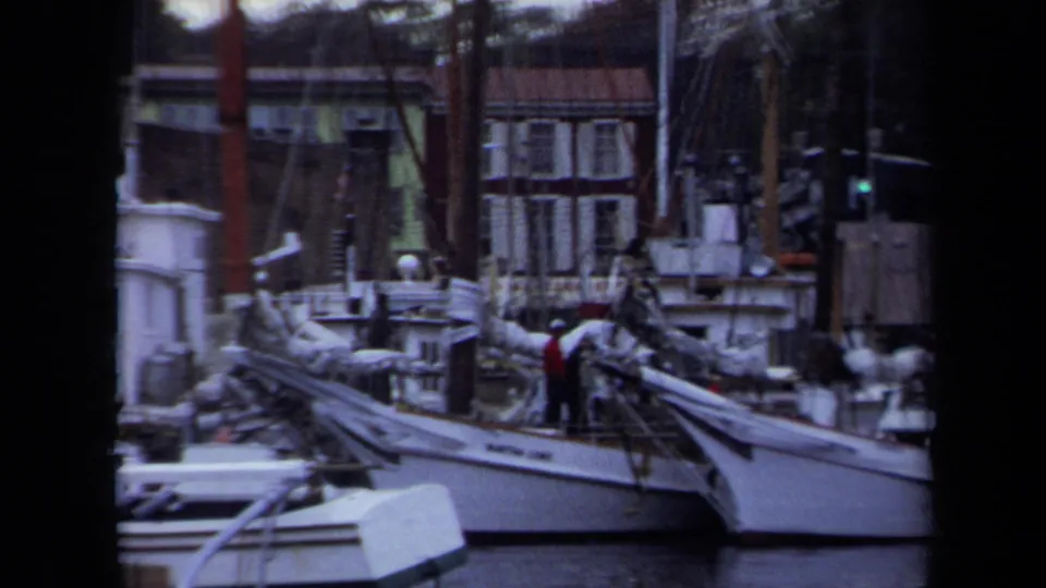 1970 Annapolis marina frame showing moored sailboats, rigging, brick waterfront buildings, and people on deck.