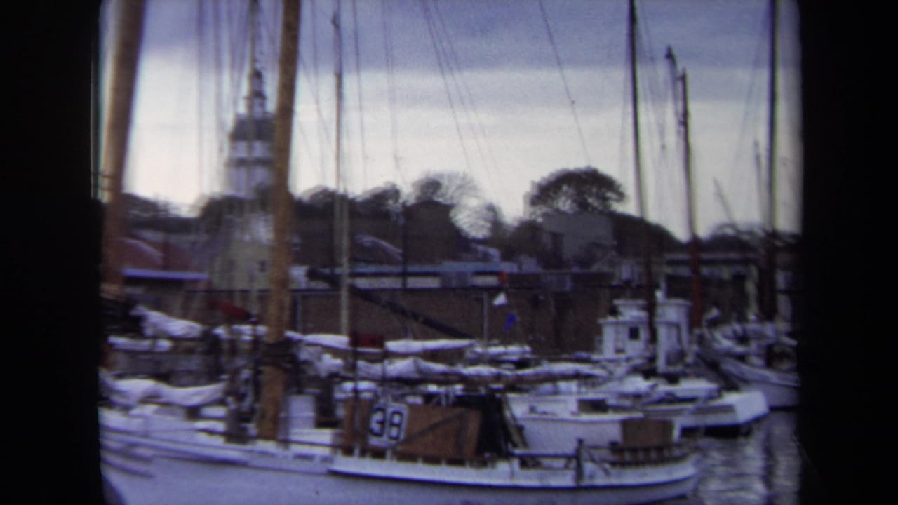 1970 Annapolis harbor view with moored sailboats, furled sails, dock structures, and a distant steeple beyond the waterfront.
