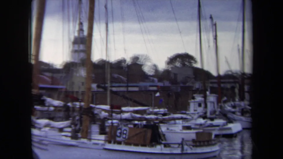 1970 Annapolis harbor view with moored sailboats, furled sails, dock structures, and a distant steeple beyond the waterfront.