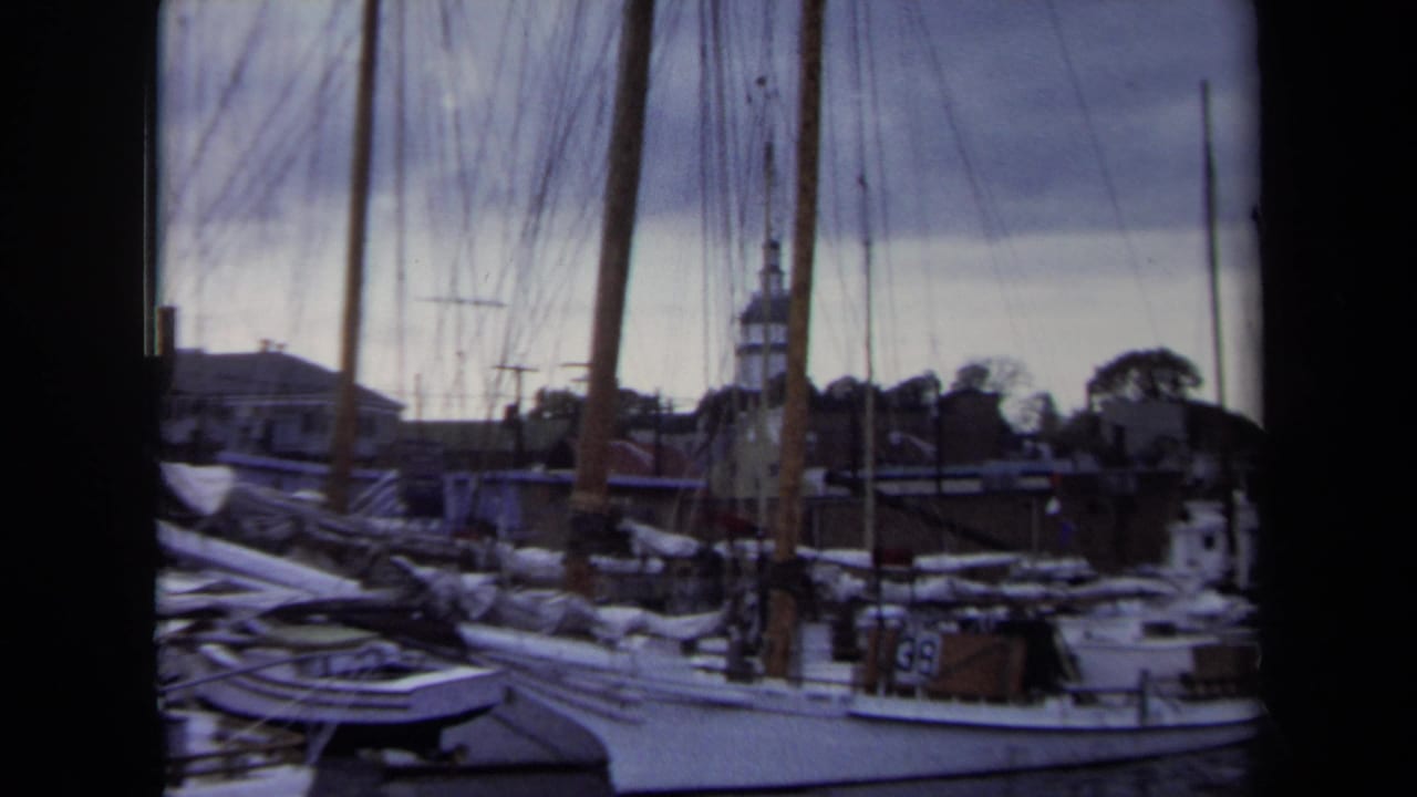 1970 Annapolis waterfront frame showing clustered sailboats, dockside rigging, water, rooftops, and a distant church-like steeple.