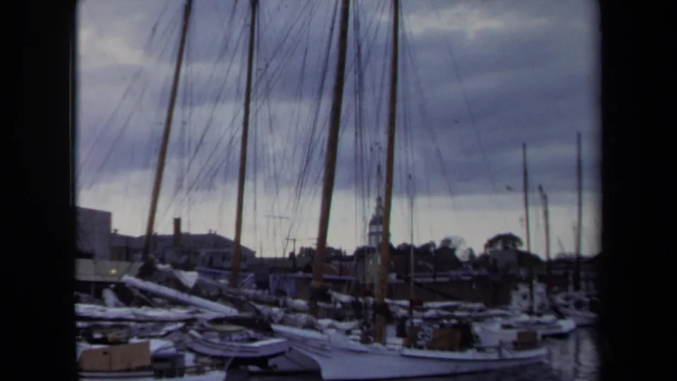 1970 Annapolis dockside view with moored sailboats, vertical masts, rigging, waterfront rooftops, and a distant steeple under cloud cover.