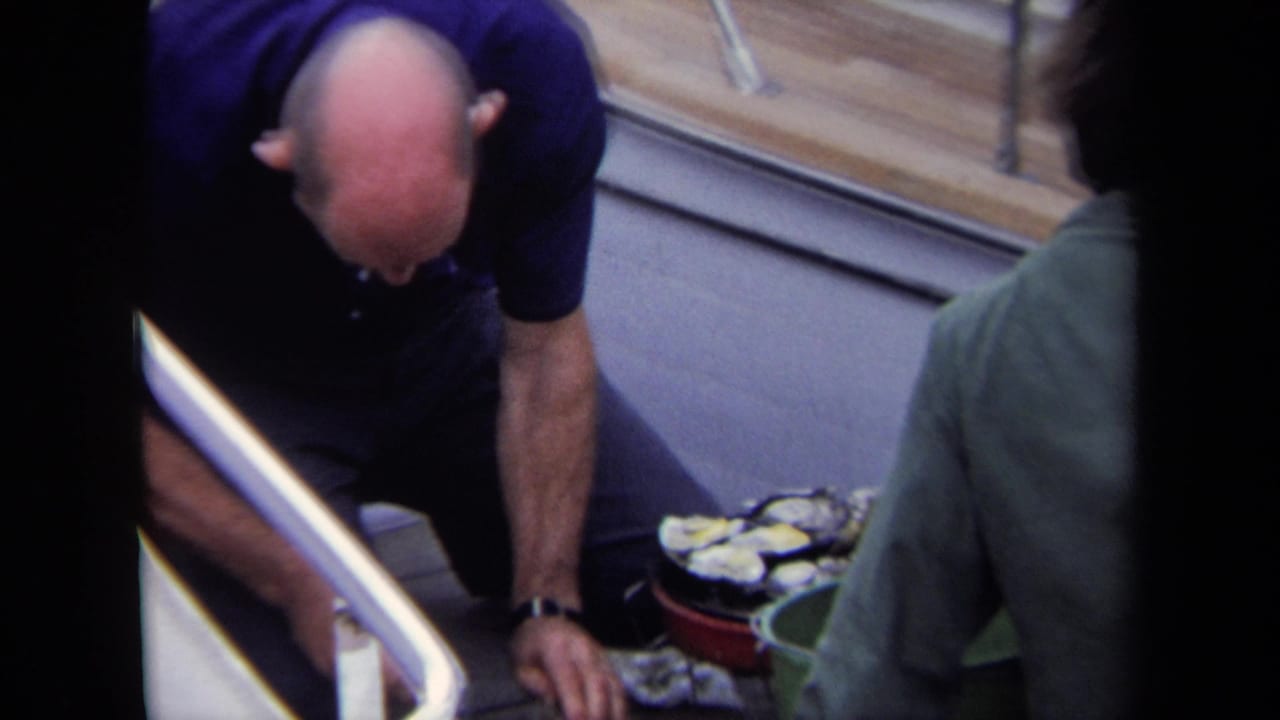 Bald man reaches toward oysters on the deck beside a man in a green jacket.