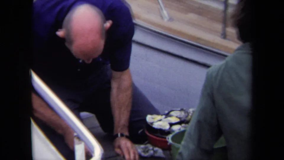 A bald man in a blue polo reaches down near oysters on the Annapolis boat deck while a man in a green jacket stands beside him.