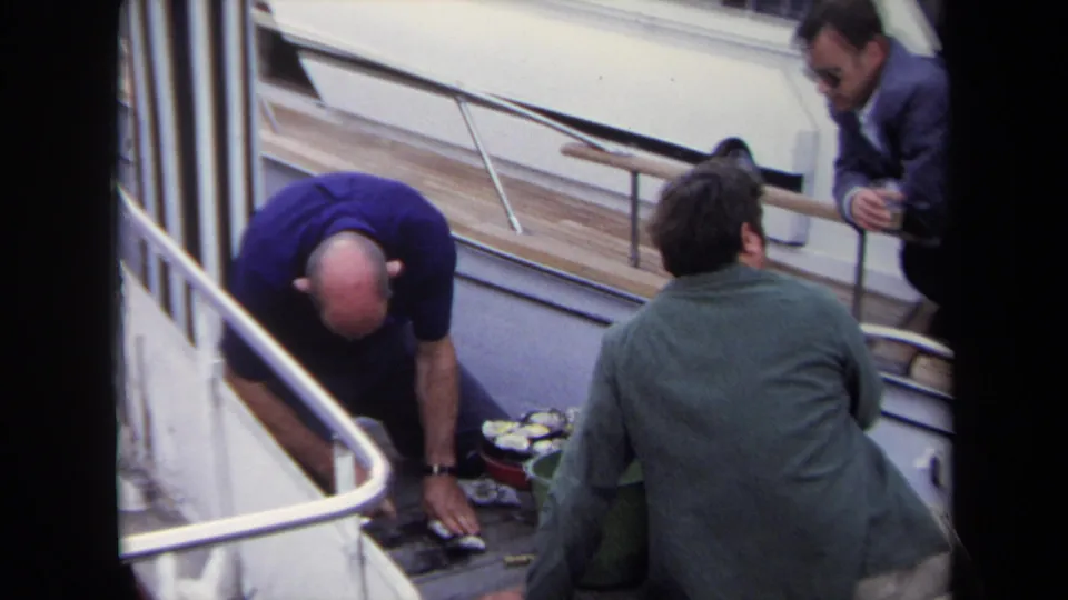 On the 1970 Annapolis boat deck, two men focus on oysters while a man in a suit and sunglasses watches from the railing.