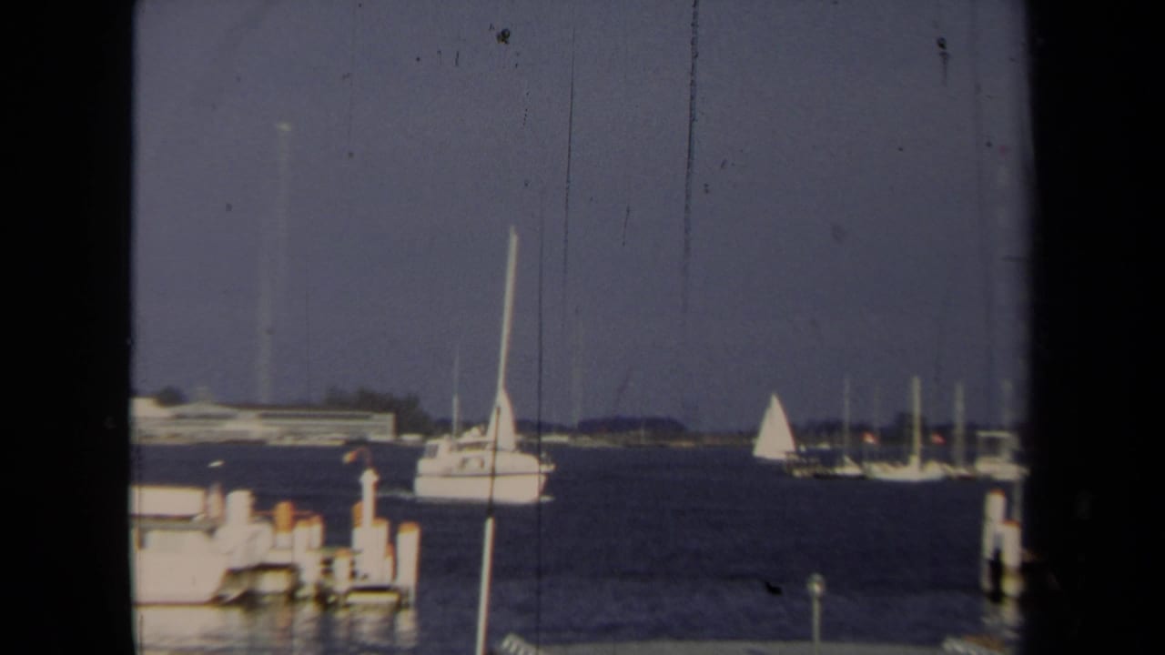 1970 Annapolis harbor view with sailboats crossing open water and docked boats near the shoreline.