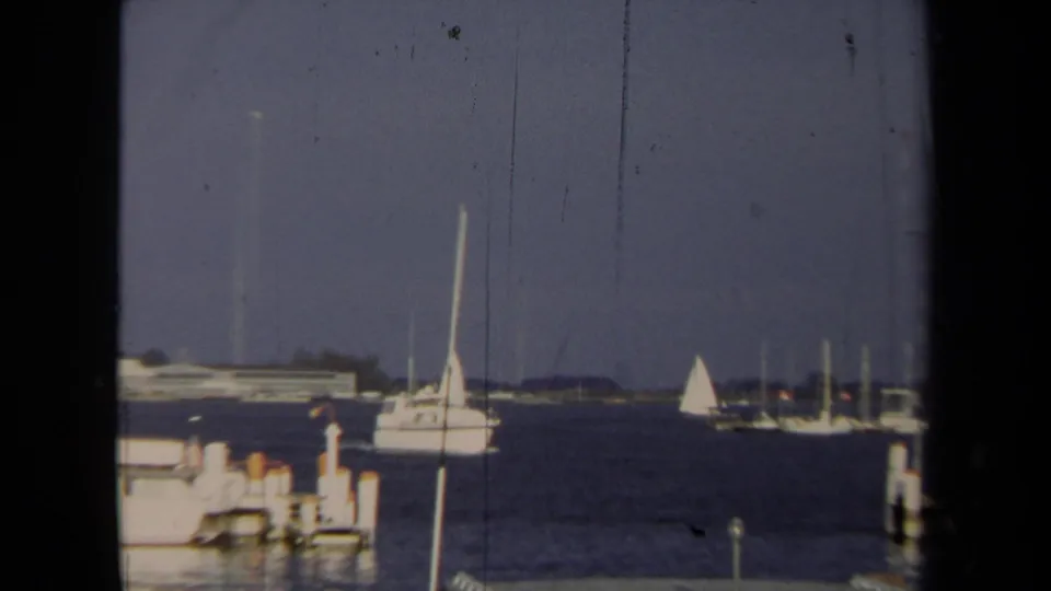 1970 Annapolis harbor view with sailboats crossing open water and docked boats near the shoreline.