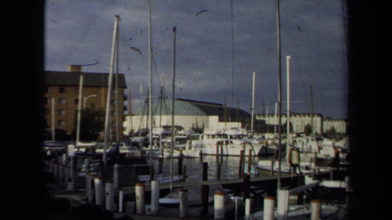 1970 Annapolis marina view with clustered masts, docked boats, pilings, and waterfront buildings under a cloudy sky.