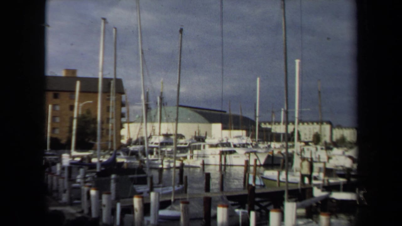 1970 Annapolis marina view with moored yachts, sailboat masts, wooden piers, water, and waterfront buildings under cloudy sky.