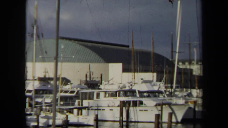 1970 Annapolis marina scene with docked yachts, sailboat masts, wooden pilings, and a large curved green-roof building behind them.