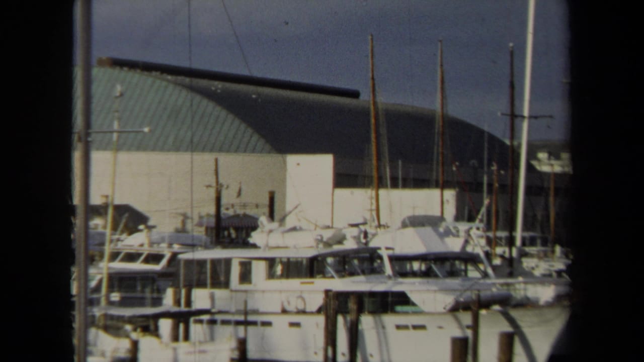 1970 Annapolis marina frame showing moored yachts, vertical masts, wooden dock posts, and the large curved green-roof building beyond.