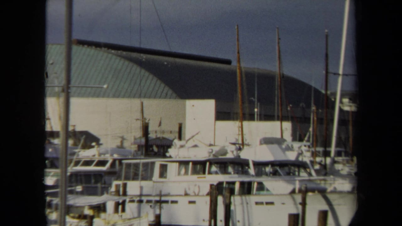 1970 Annapolis marina view with moored cabin boats, sailboat masts, wooden pilings, and a large curved-roof building behind the docks.