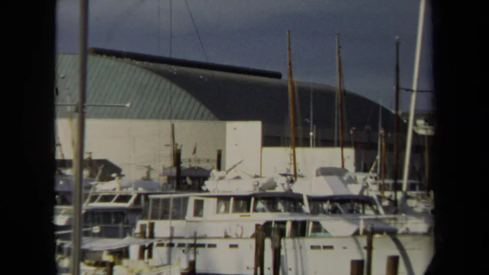 1970 Annapolis marina view with moored cabin boats, sailboat masts, wooden pilings, and a large curved-roof building behind the docks.