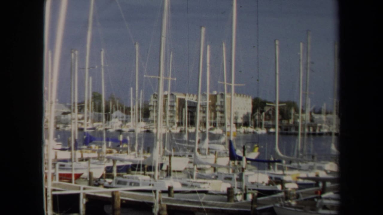1970 Annapolis 8mm frame of a marina foreground with boats, tall masts, wooden posts, water, and waterfront buildings in soft focus.