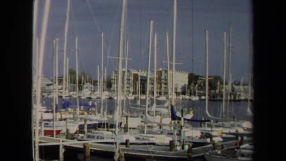 1970 Annapolis 8mm frame of a marina foreground with boats, tall masts, wooden posts, water, and waterfront buildings in soft focus.