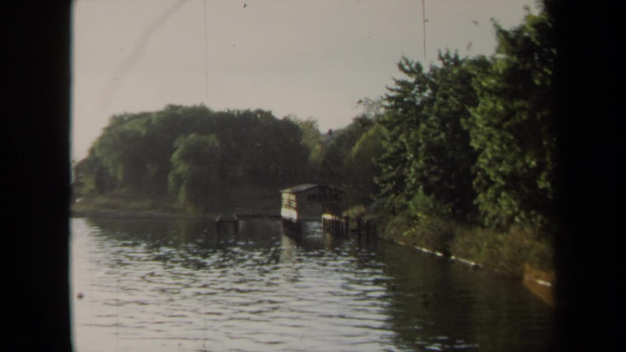 1970 Annapolis shoreline view with a calm waterway, dense green trees, a small boathouse-like structure, and ripples across the water.