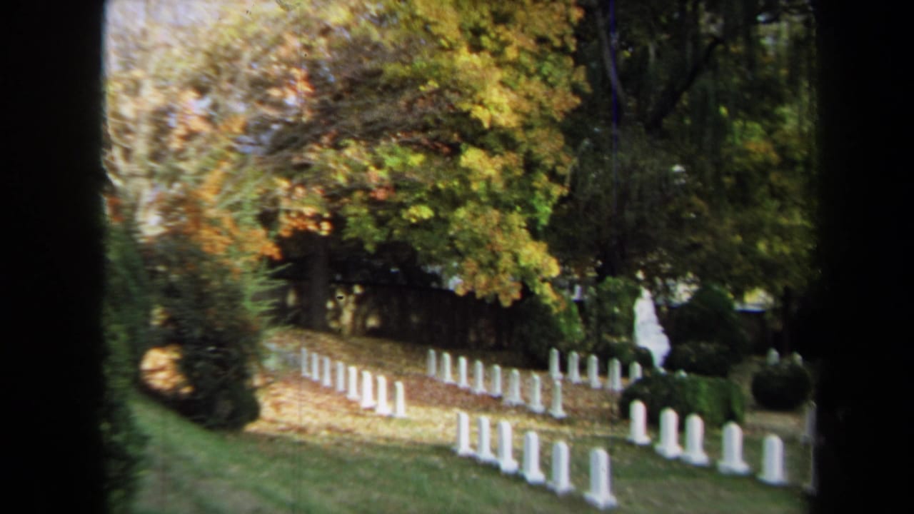 1970 Annapolis 8mm frame of cemetery headstones on a slope, framed by trees and a distant visitor.