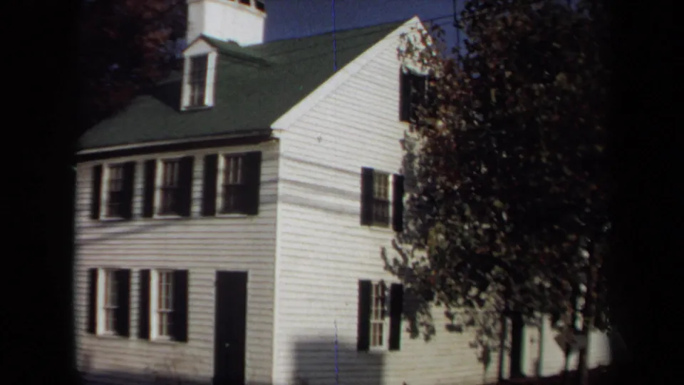 1970 Annapolis home-movie frame of a two-story white clapboard house with black shutters, green roof, dark door, and leafy tree.