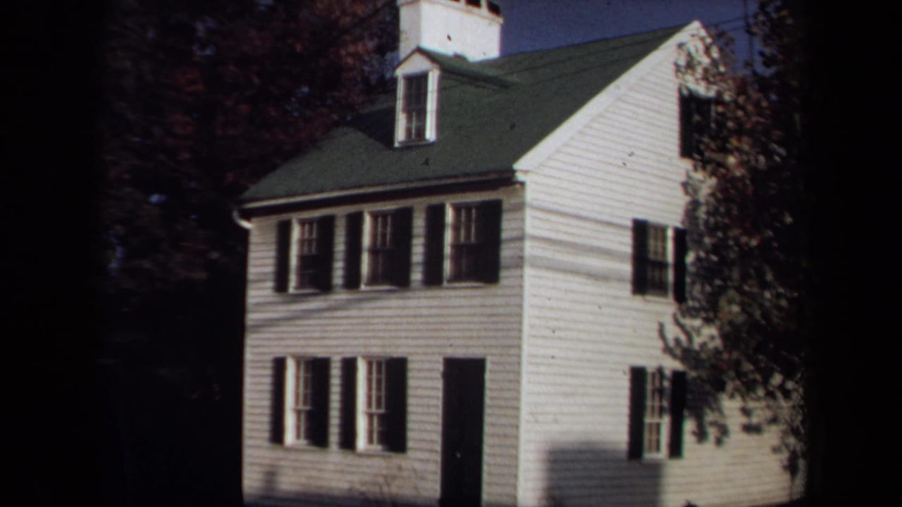 1970 Annapolis home-movie frame showing a white colonial-style house with dark shutters, green roof, dormer and surrounding trees.