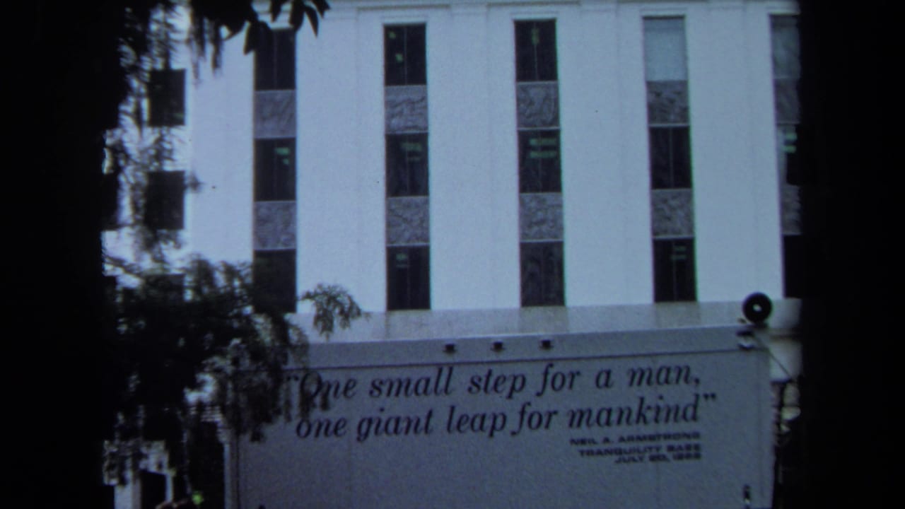 1970 California 8mm frame showing a white building, dark foreground trees, and a public banner with Neil Armstrong's moon-landing quote.