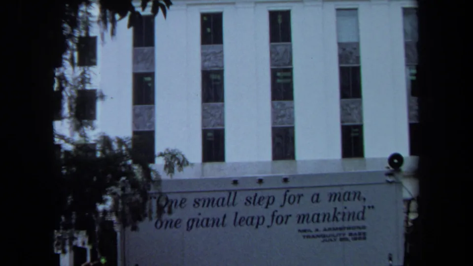 1970 California 8mm frame showing a white building, dark foreground trees, and a public banner with Neil Armstrong's moon-landing quote.