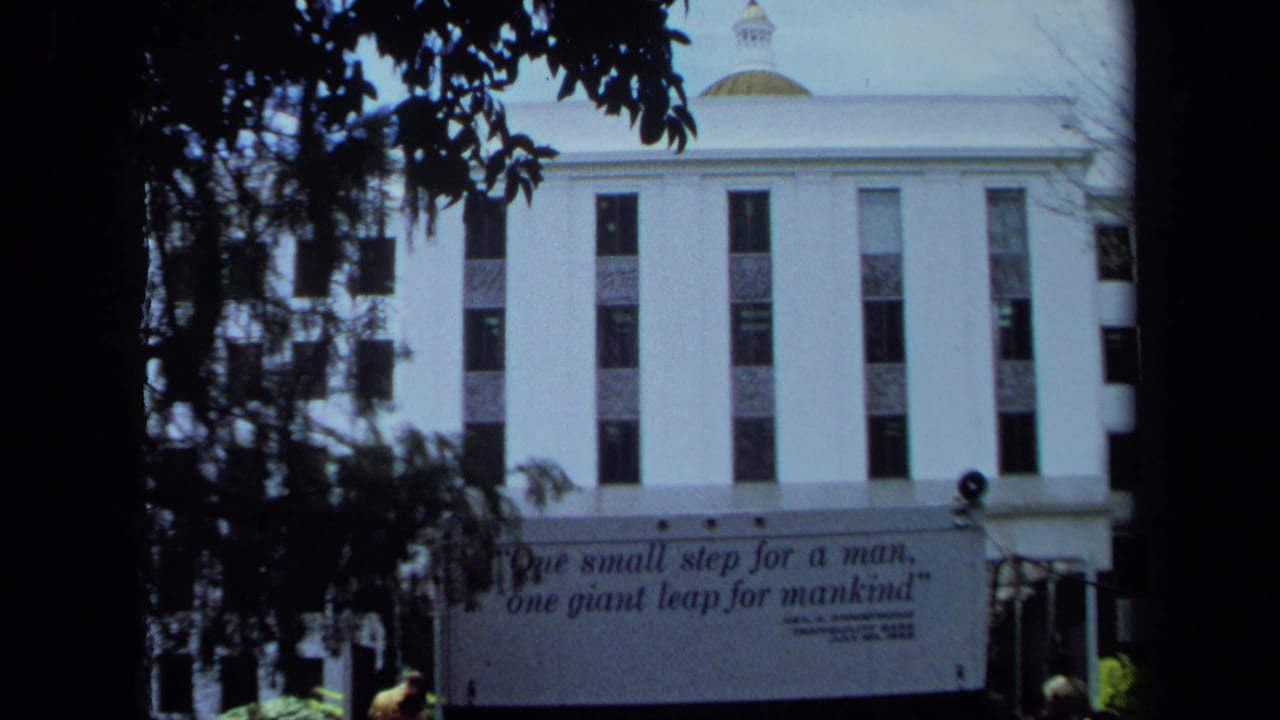 1970 California home-movie frame with tree branches framing a white building and a partially visible Armstrong quote banner.