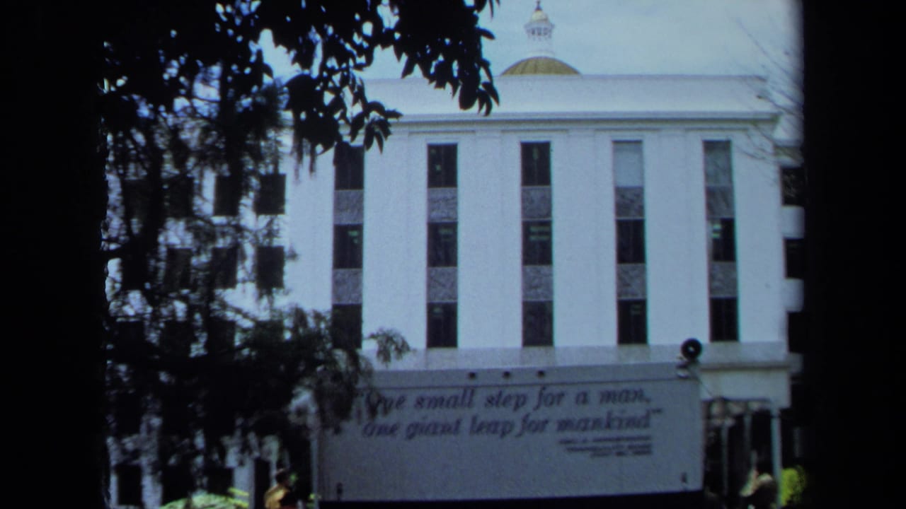 1970 California 8mm frame showing the Armstrong quote banner across a white building, seen through shaded trees.