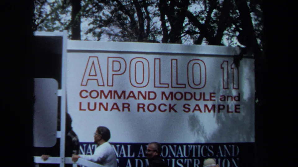 1970 California 8mm frame showing visitors beside a large Apollo 11 command module and lunar rock sample exhibition sign.