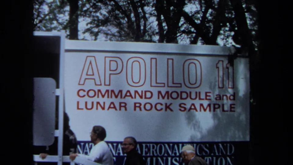 Visitors in a 1970 California home movie move past the Apollo 11 command module and lunar rock sample sign in daylight.