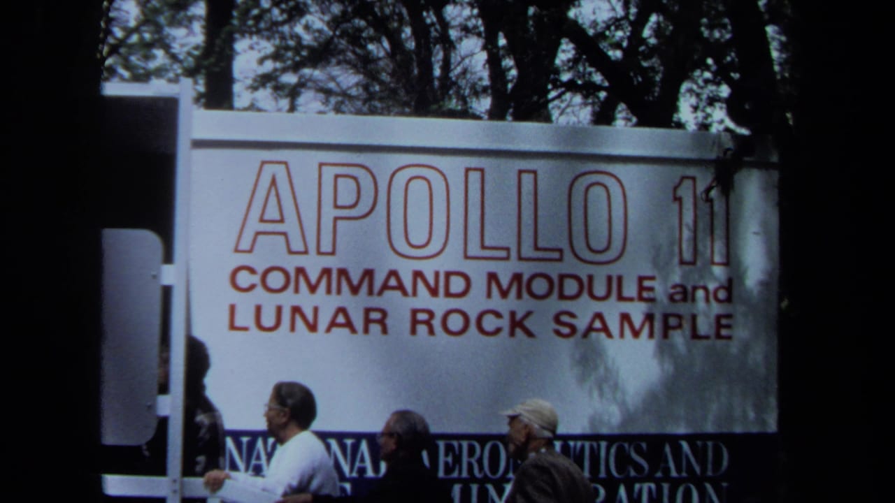 A handheld 8mm view shows several visitors beneath the Apollo 11 command module and lunar rock sample display board.