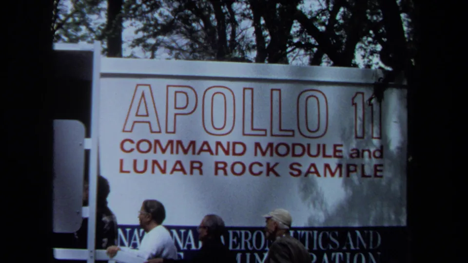 A handheld 8mm view shows several visitors beneath the Apollo 11 command module and lunar rock sample display board.