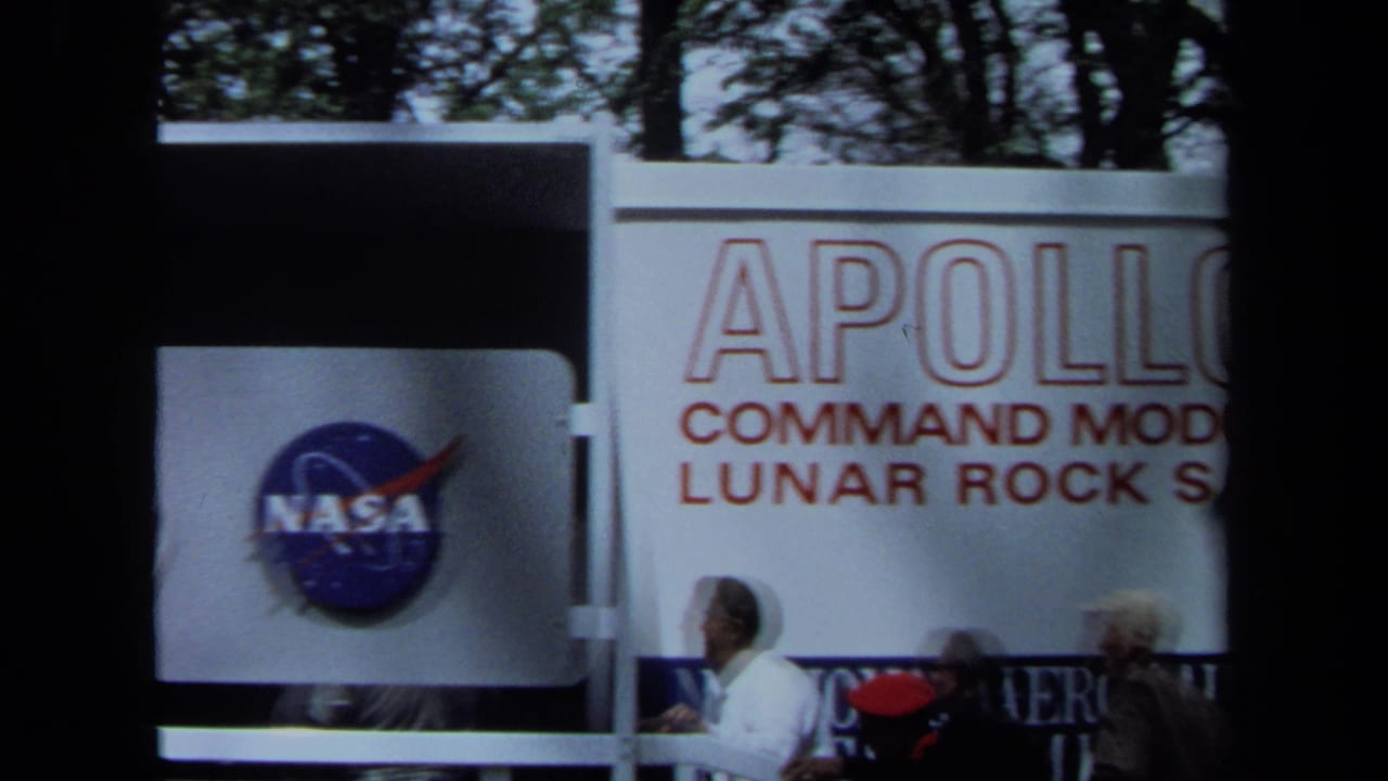 The frame shows a NASA emblem on the exhibit structure with visitors and partial Apollo 11 display text nearby.