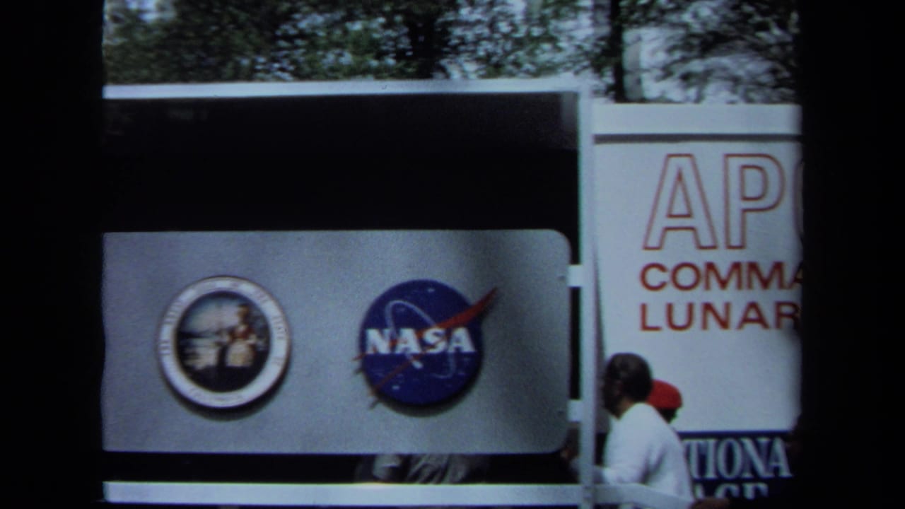 1970 California 8mm frame showing NASA and Apollo mission emblems on an exhibit panel, with partial Apollo command and lunar room text behind.