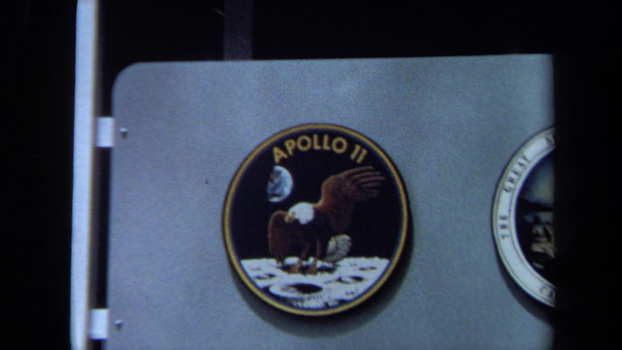 Close 1970 California 8mm view of a large Apollo emblem on a display panel, with another round mission patch partly cut off at right.