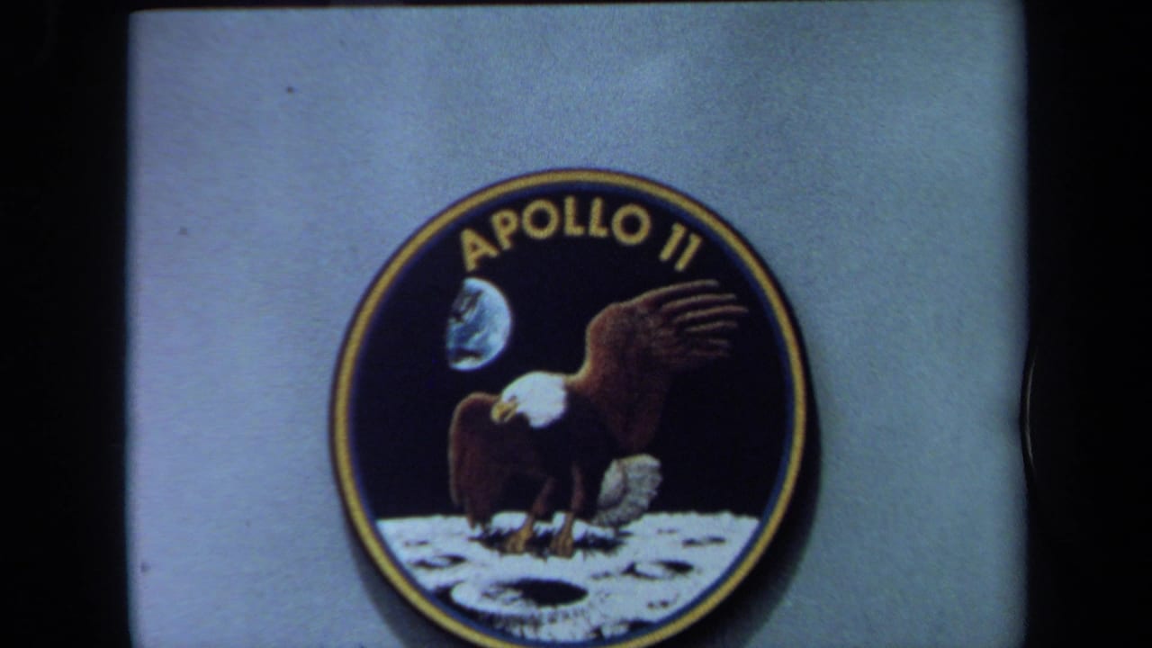 1970 California 8mm frame centered on a large Apollo emblem mounted to a pale exhibit panel, with soft grain and dark edge framing.