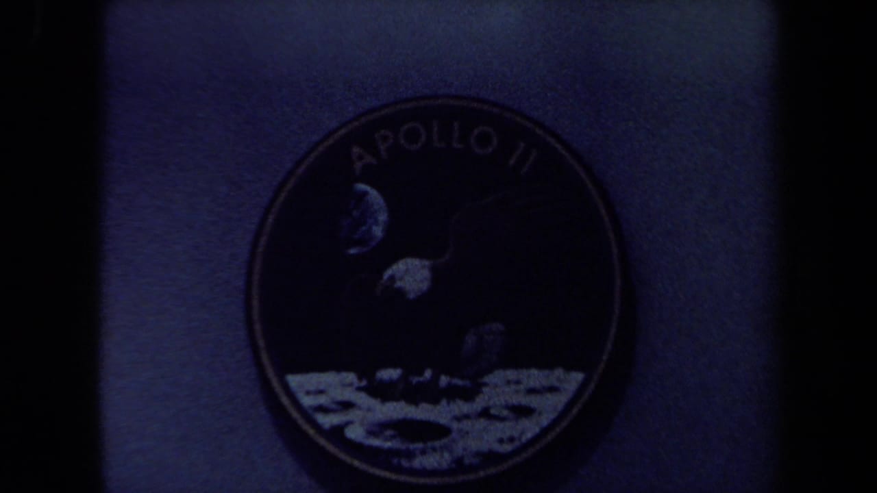 Underexposed 8mm frame showing the Apollo 11 patch in shadow, with the eagle and lunar artwork partly visible.