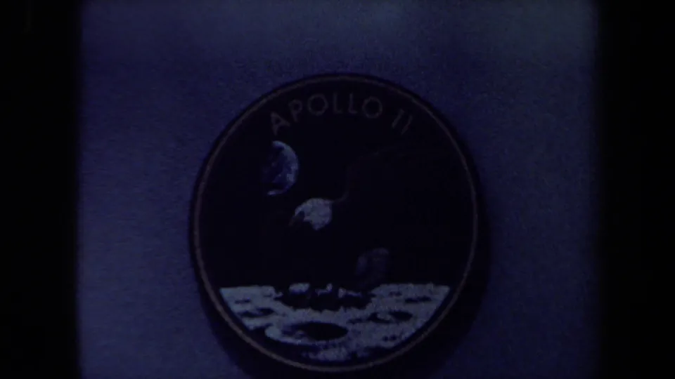 Underexposed 8mm frame showing the Apollo 11 patch in shadow, with the eagle and lunar artwork partly visible.