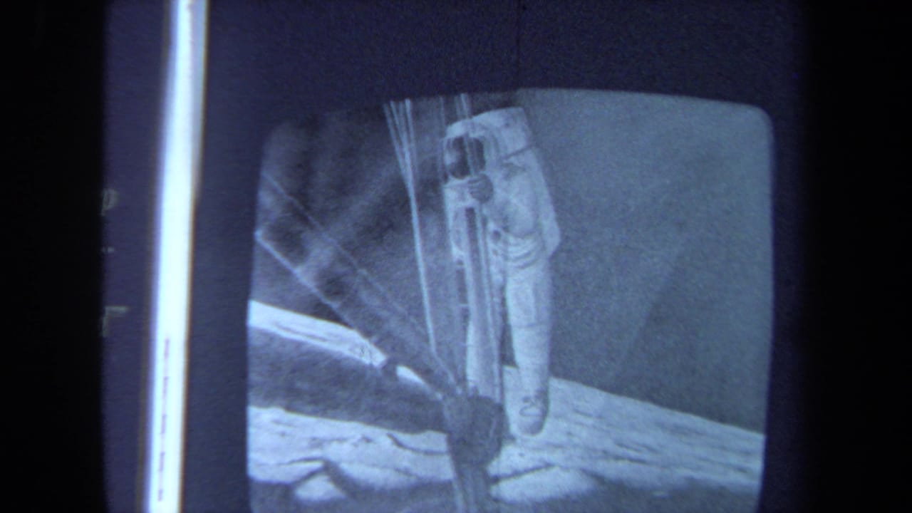Grainy 8mm frame of monochrome moonwalk footage, showing an astronaut on the lunar surface beside equipment-like forms.