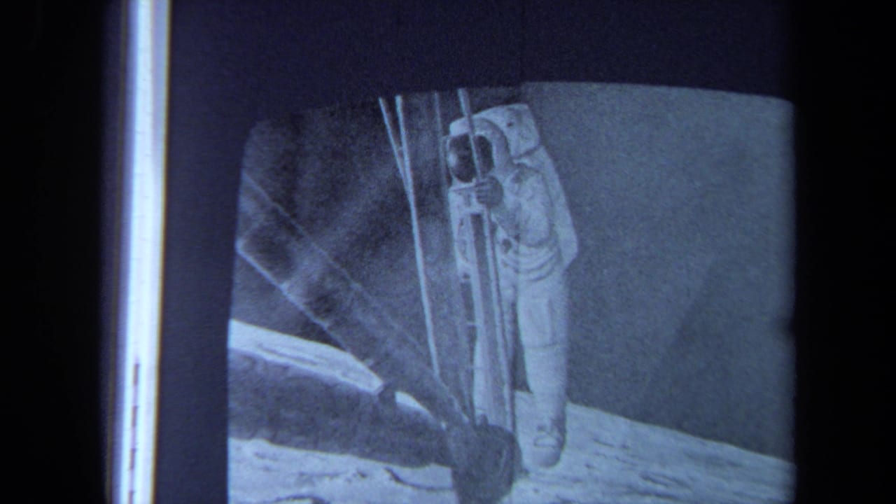 1970 California 8mm frame of a filmed television screen showing an astronaut in a white suit near a ladder on a lunar-looking surface.
