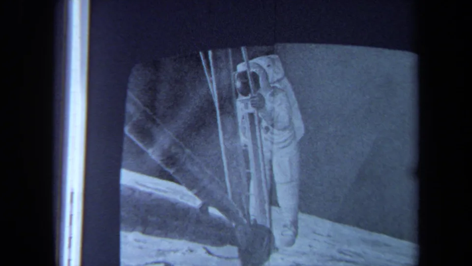1970 California 8mm frame of a filmed television screen showing an astronaut in a white suit near a ladder on a lunar-looking surface.