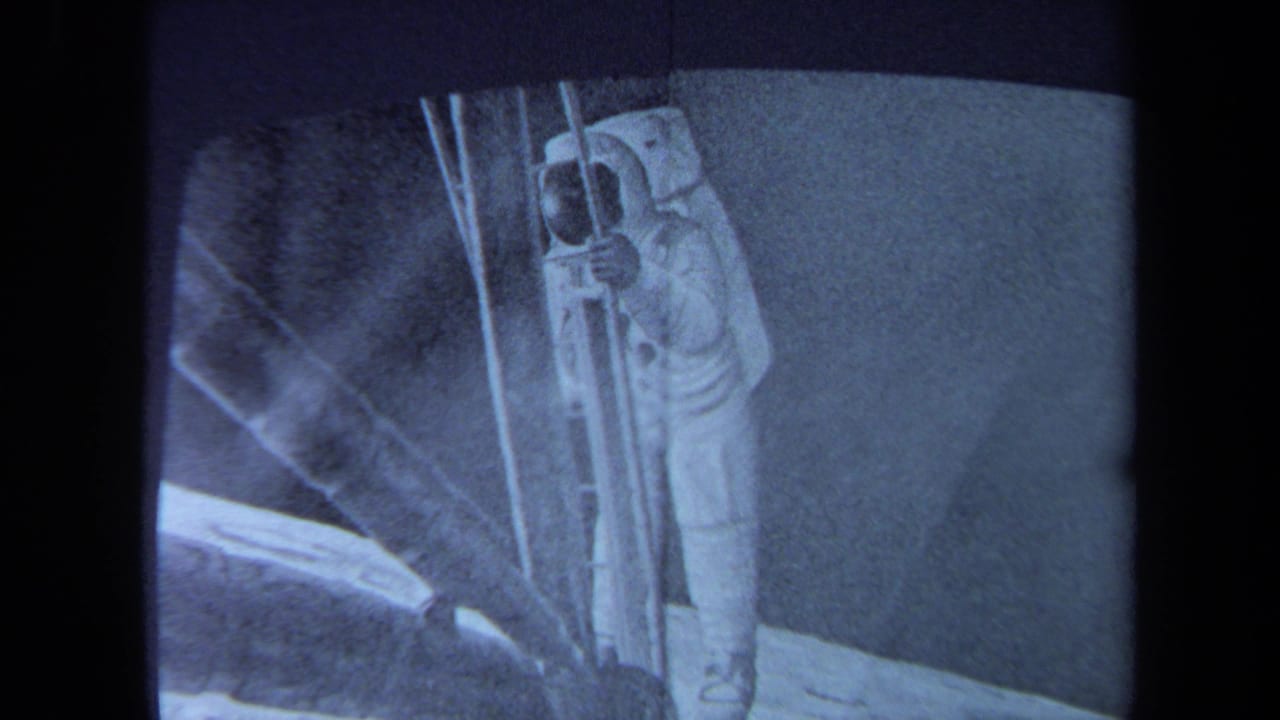 A closer 8mm view of the television image centers the astronaut, ladder-like equipment, and pale lunar-looking ground.