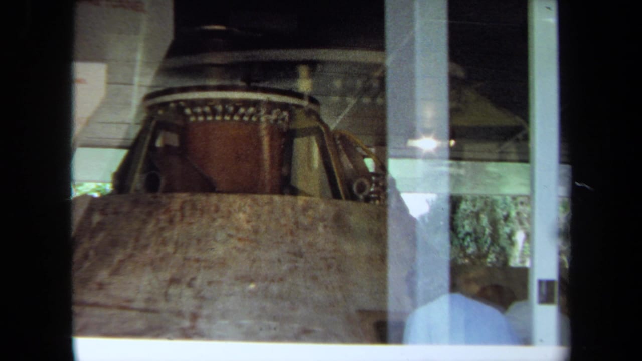 Wider 1970 California 8mm view of the float form indoors, seen through glass with reflections and a partially visible person below.