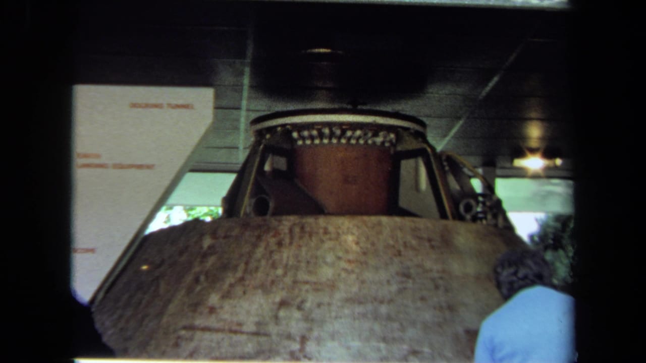 1970 California 8mm frame showing visitors near a weathered cone-shaped spacecraft module with exhibit signage behind it.