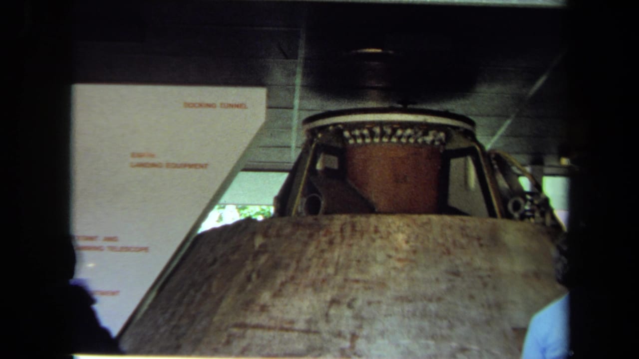Space capsule exhibit seen from behind visitors, with signs in the background.